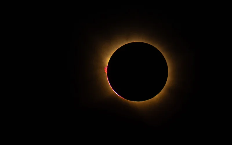 Un eclipse solar total con la corona brillante y prominencias rojas visibles en los bordes.