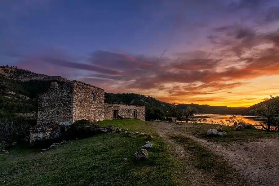 Ruinas de piedra antigua junto a un lago al atardecer en el Matarraña, con colinas y cielo en tonos púrpura y naranja
