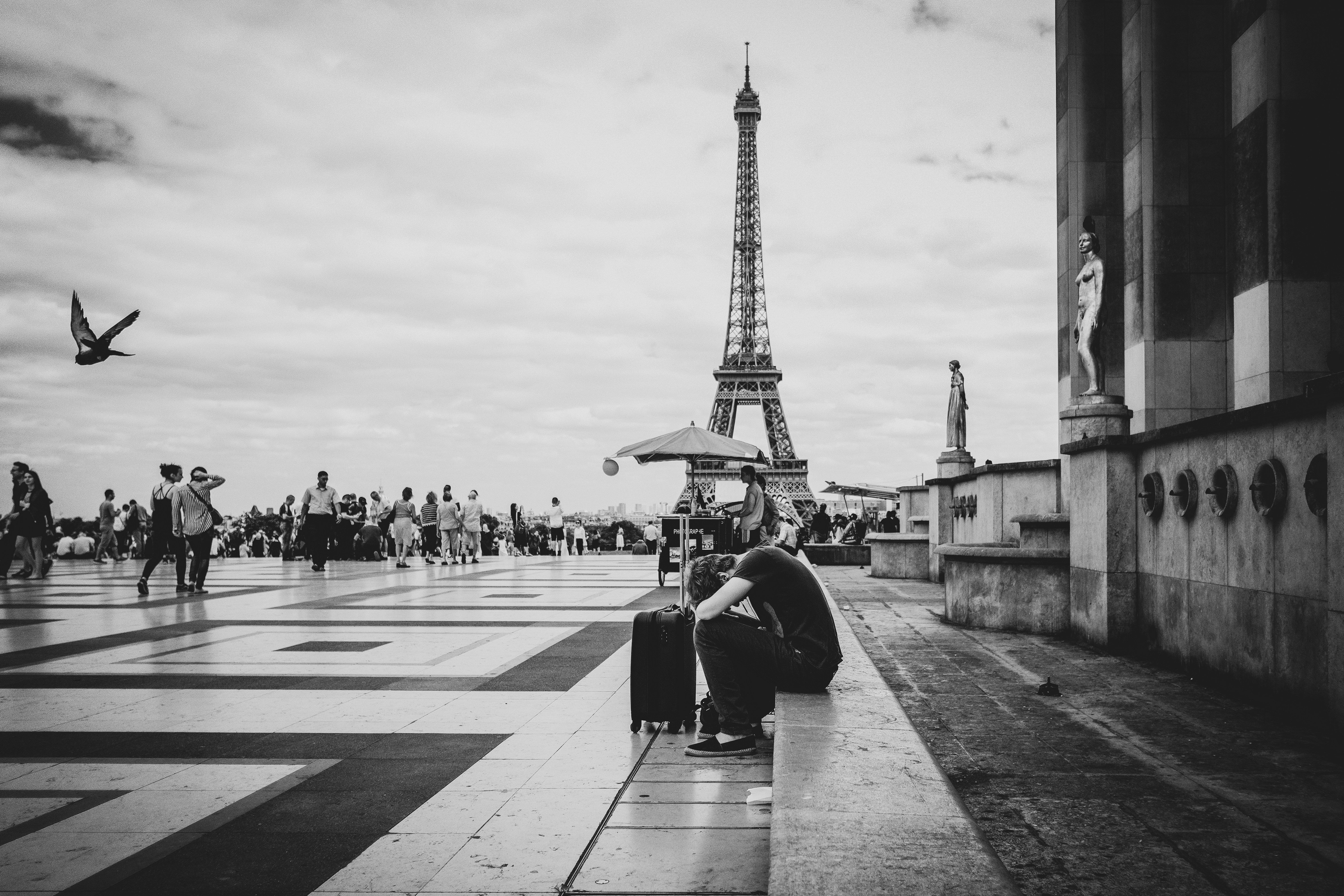 Hombre sentado con maleta frente a la Torre Eiffel y gente caminando, en blanco y negro.