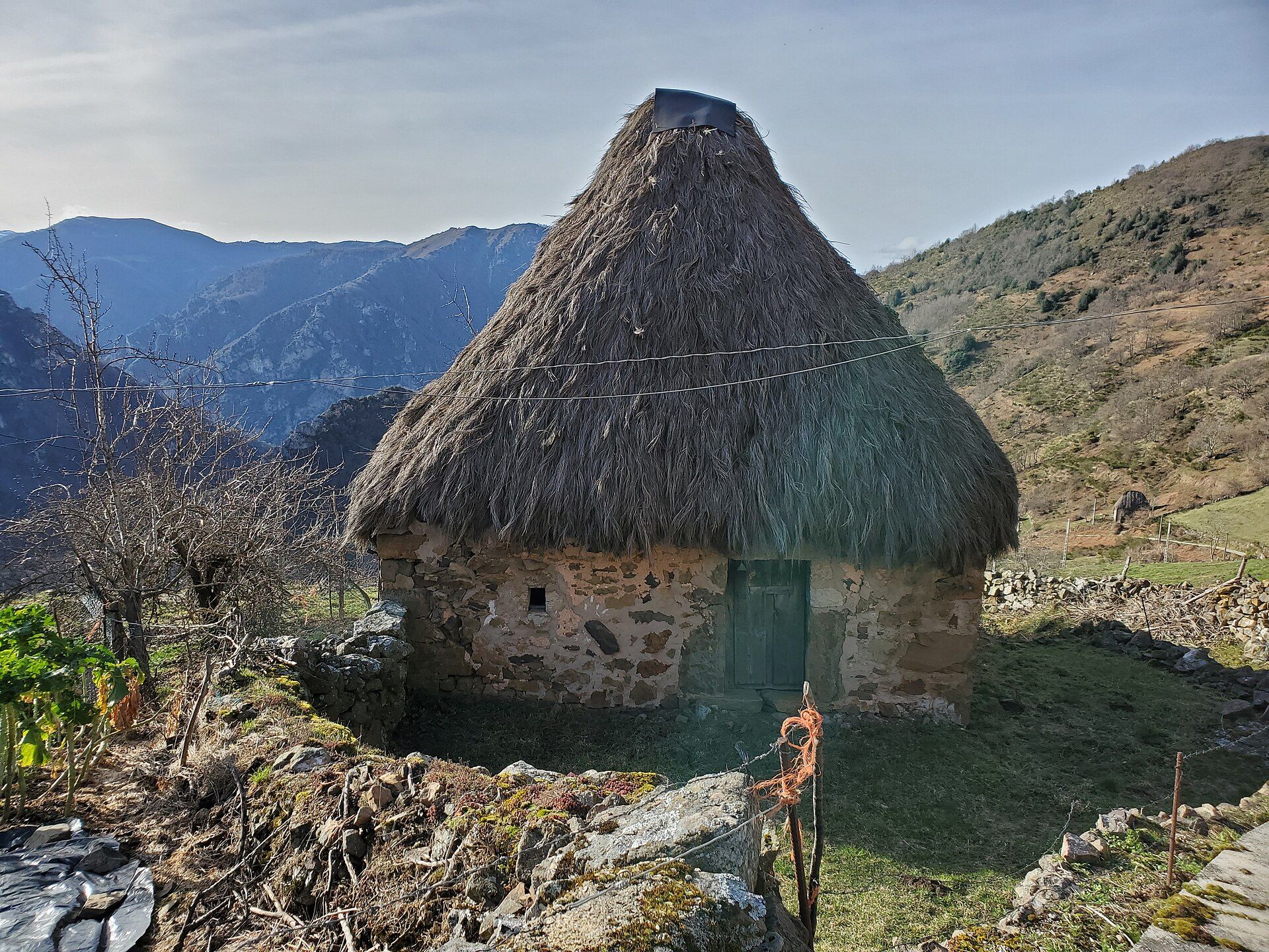 Un teito tradicional de piedra con tejado de paja en un paisaje montañoso soleado.