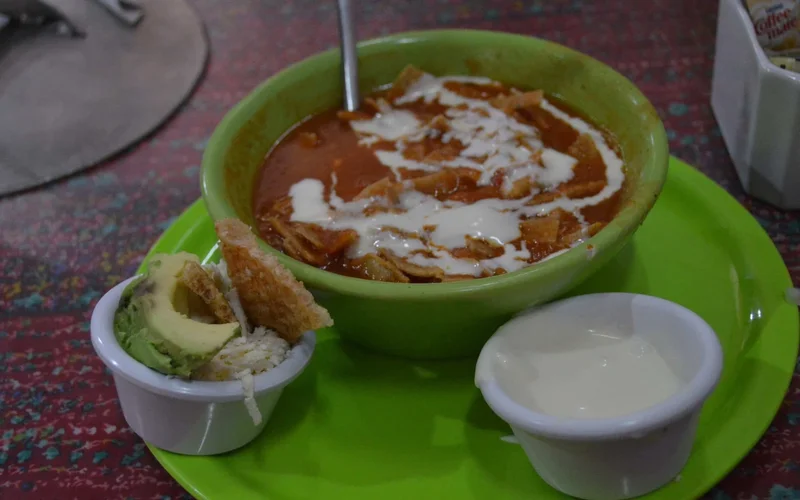 Un tazón verde con guiso rojo cubierto de crema blanca y chips de tortilla, junto a recipientes de aguacate y crema sobre un plato verde en una mesa