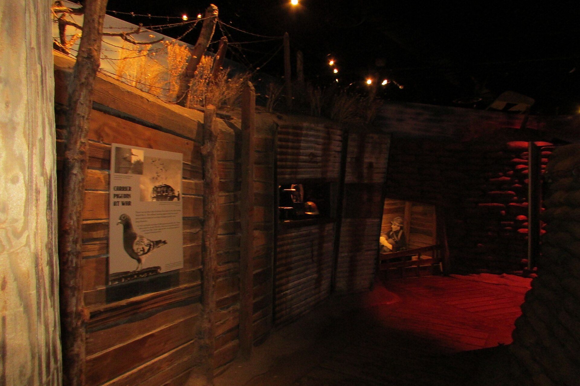 Interior de una recreación de trinchera oscura con paredes de madera, alambre de púas y un cartel informativo.