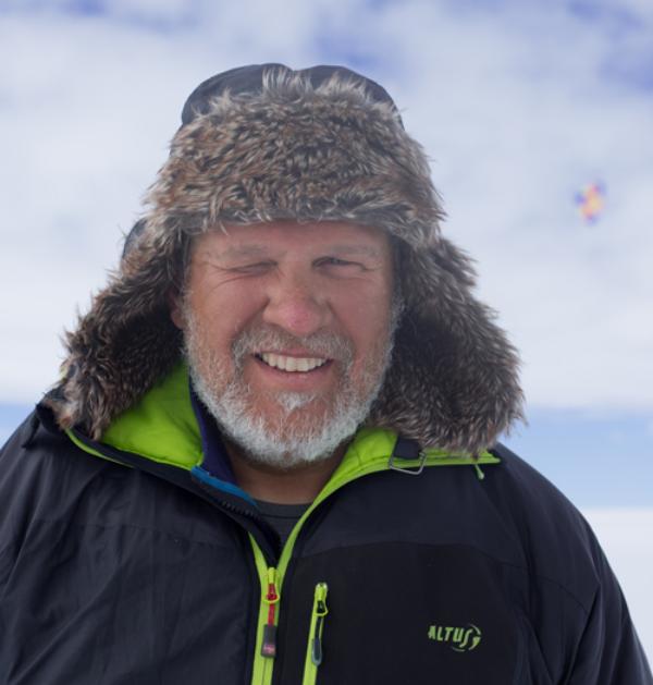 Retrato de Ramón Hernando de Larramendi, explorador sonriente con barba blanca, gorro de piel y chaqueta negra en paisaje ártico nevado