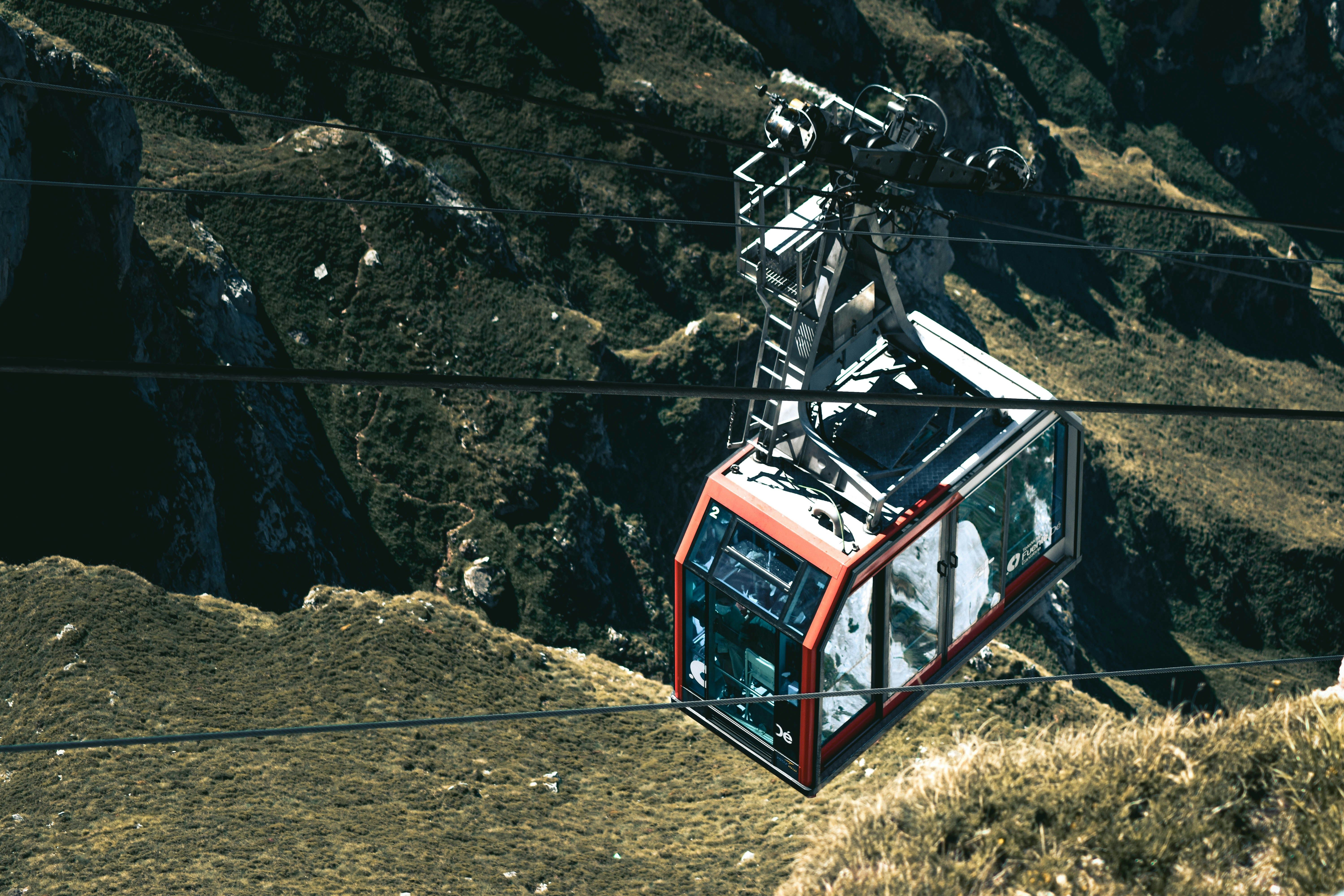 Teleférico rojo y gris suspendido sobre un paisaje montañoso verde y rocoso bajo el sol.
