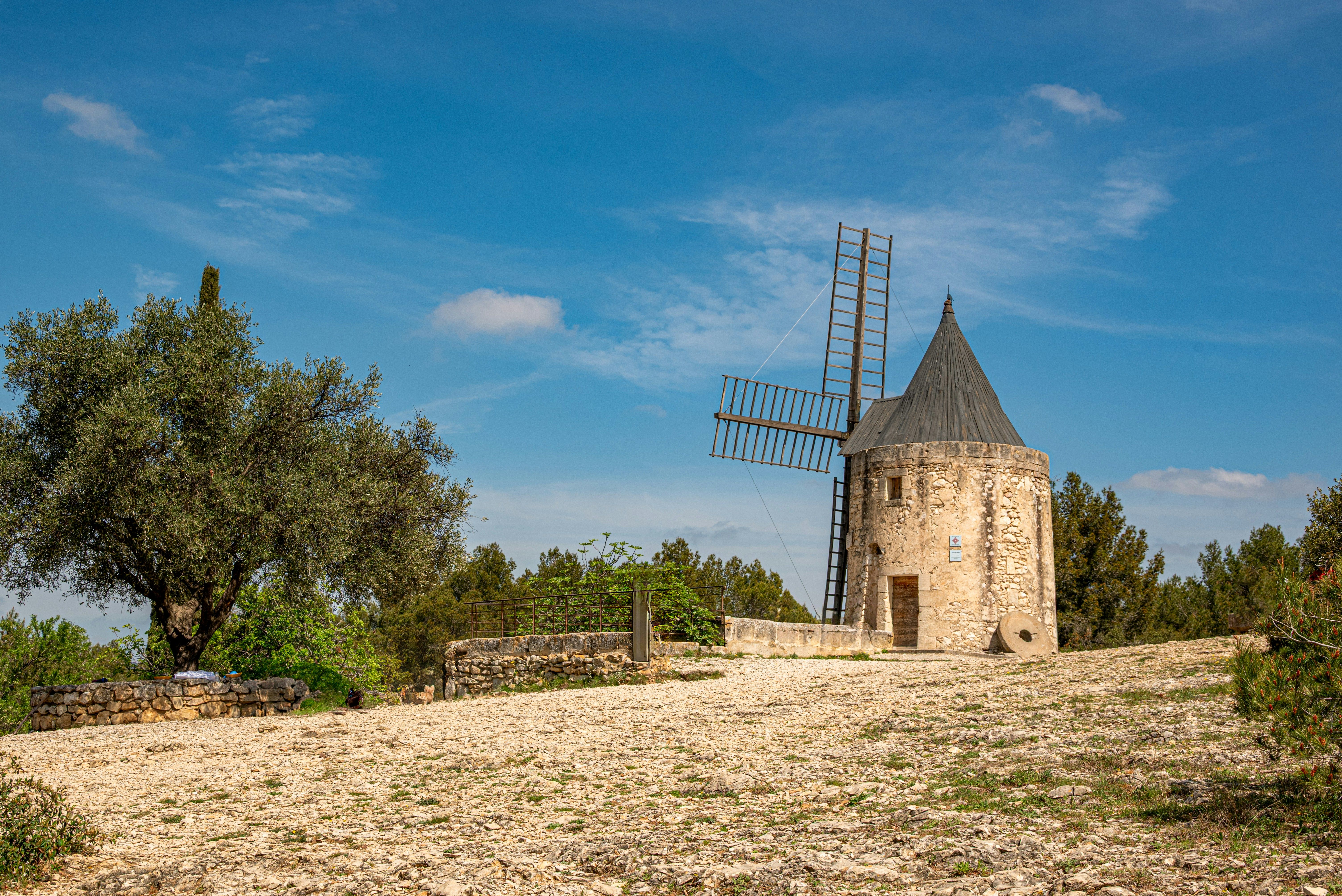 Molino de viento de piedra con tejado cónico y aspas de madera bajo un cielo azul claro.