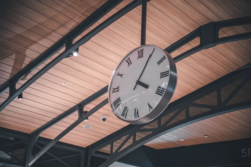 Un reloj de pared grande con números romanos cuelga del techo de madera y vigas metálicas.