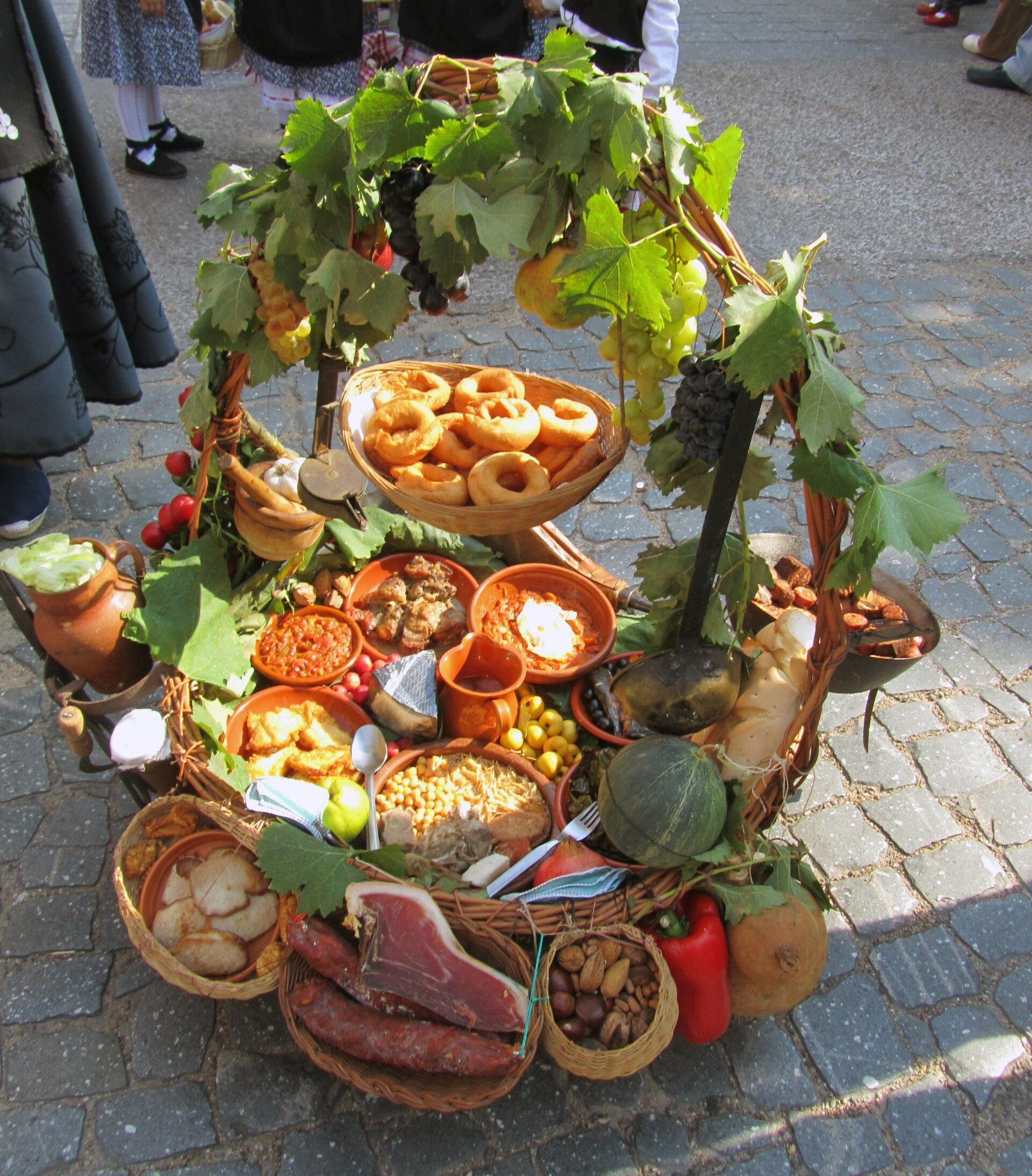 Cesta grande con uvas, hojas de parra, embutidos, platos de comida tradicional y dulces en un suelo empedrado.