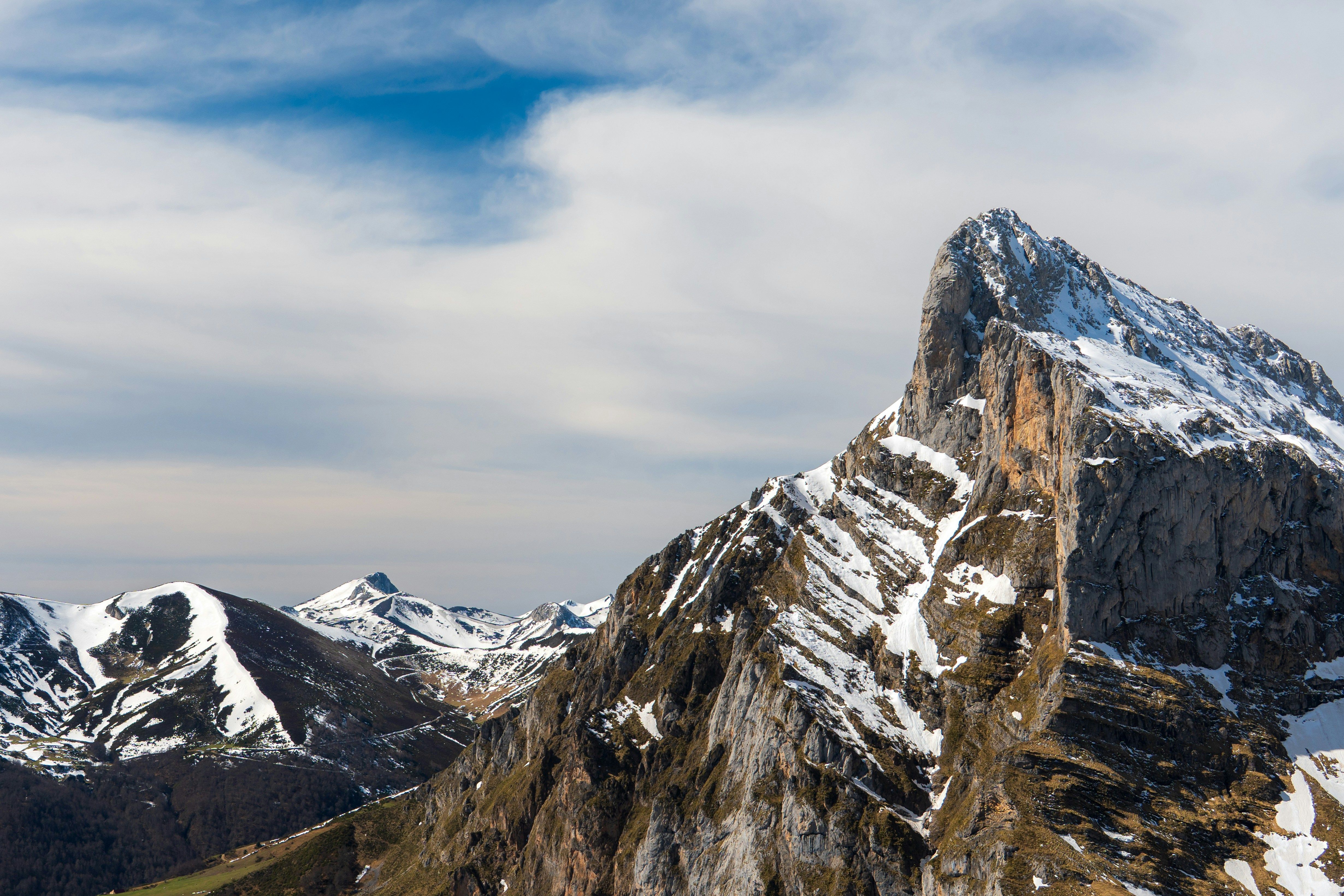 Cima de montaña rocosa cubierta de nieve bajo un cielo parcialmente nublado con otras montañas al fondo.