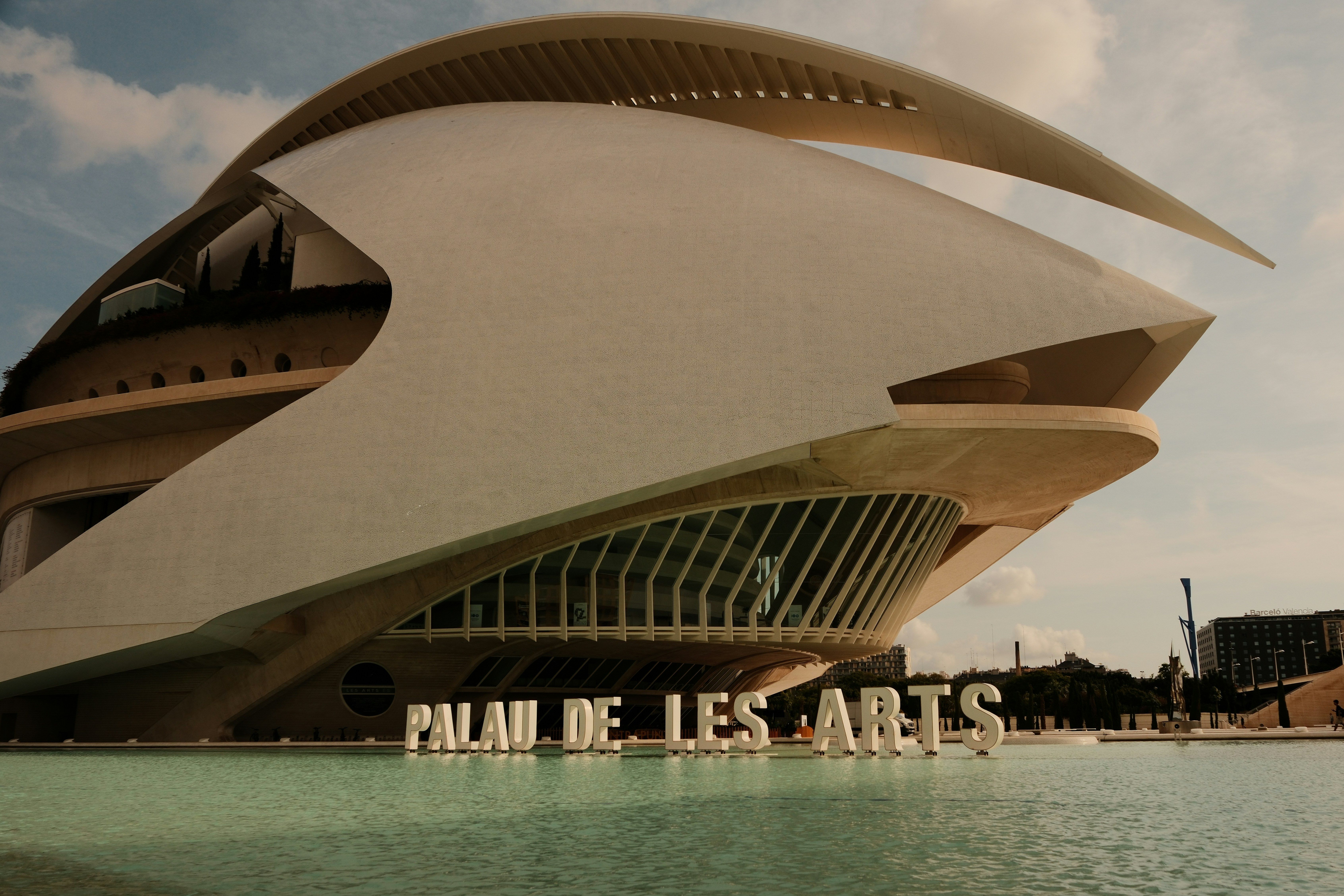El Palau de les Arts en Valencia, edificio futurista con techo curvo blanco y formas geométricas, reflejado en el agua bajo un cielo parcialmente