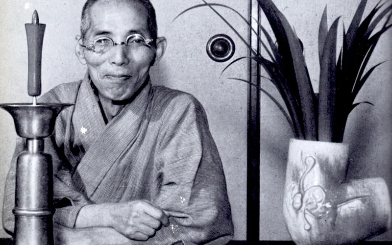 Retrato en blanco y negro de Shōji Hamada, sentado con kimono tradicional ante una mesa con vela, recipiente y planta en maceta de cerámica