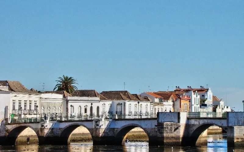 Puente de piedra con arcos sobre el río Gilão en Tavira, con edificios blancos y coloridos a las orillas, palmeras y cielo azul