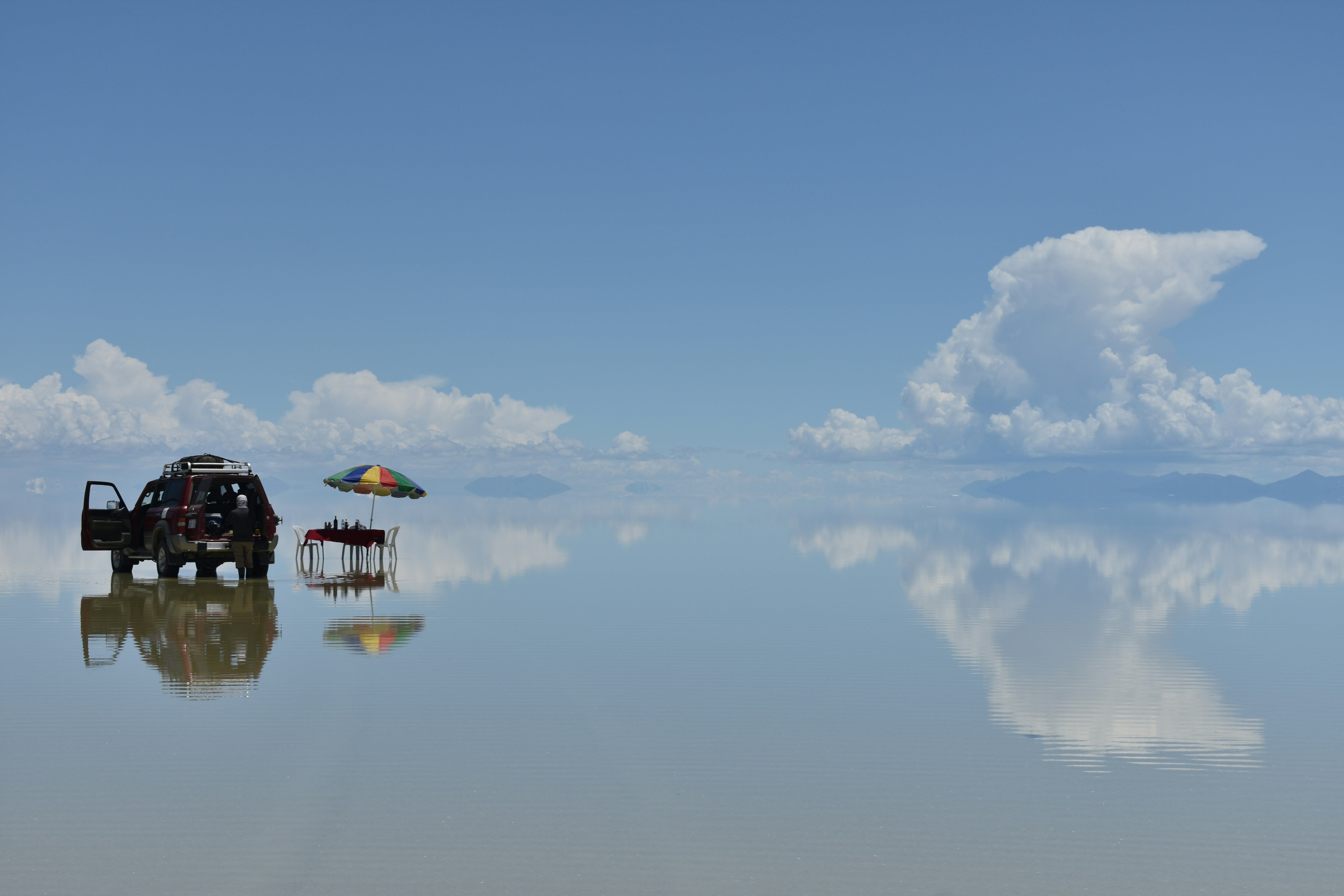 Un vehículo todoterreno y una mesa con sombrilla reflejados en una superficie de agua que refleja el cielo.