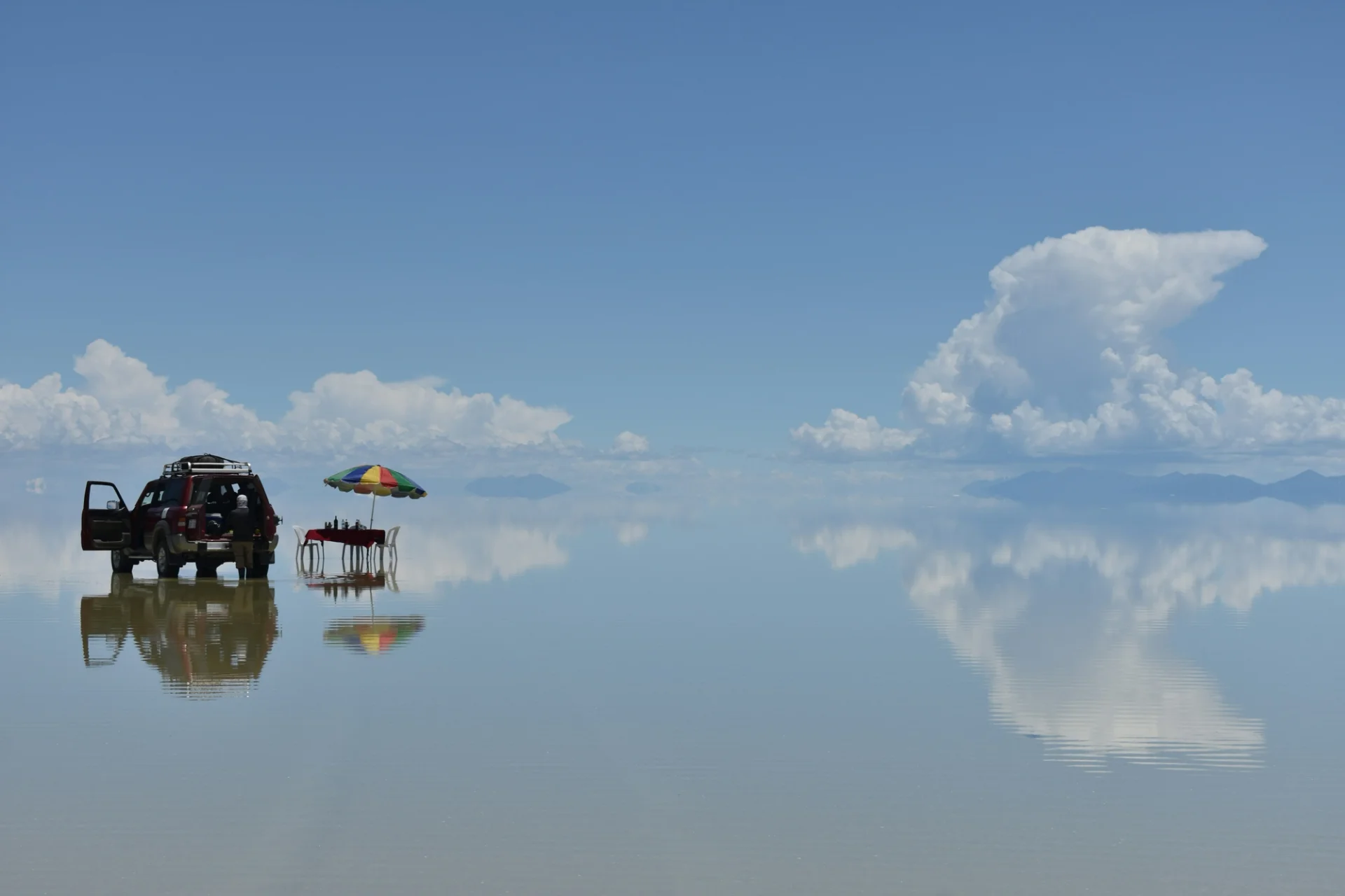 Un vehículo todoterreno y una mesa con sombrilla reflejados en una superficie de agua que refleja el cielo.