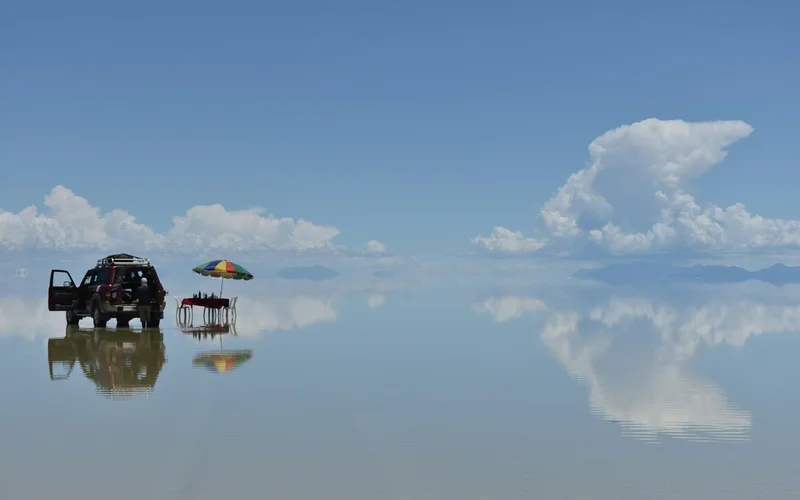 Un vehículo todoterreno y una mesa con sombrilla reflejados en una superficie de agua que refleja el cielo.
