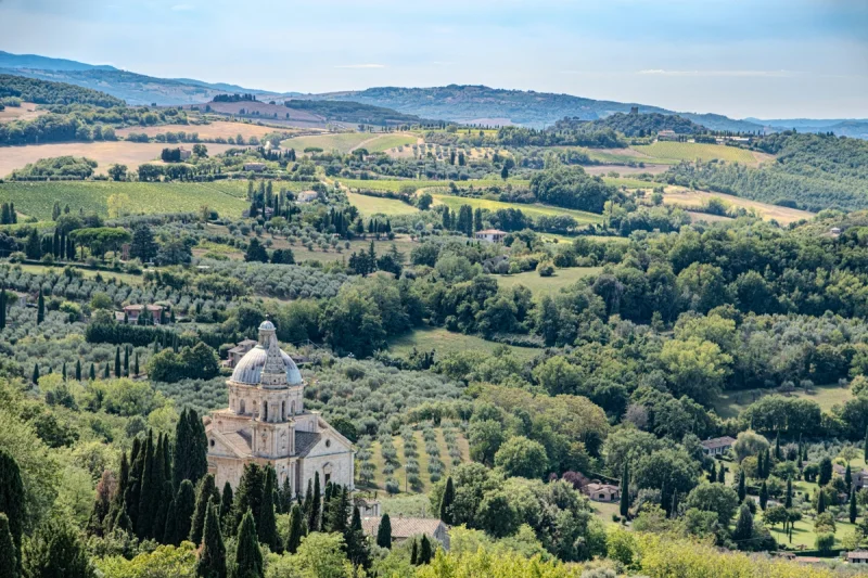 Vista aérea de una iglesia con cúpula entre cipreses y campos verdes ondulados en la Toscana.