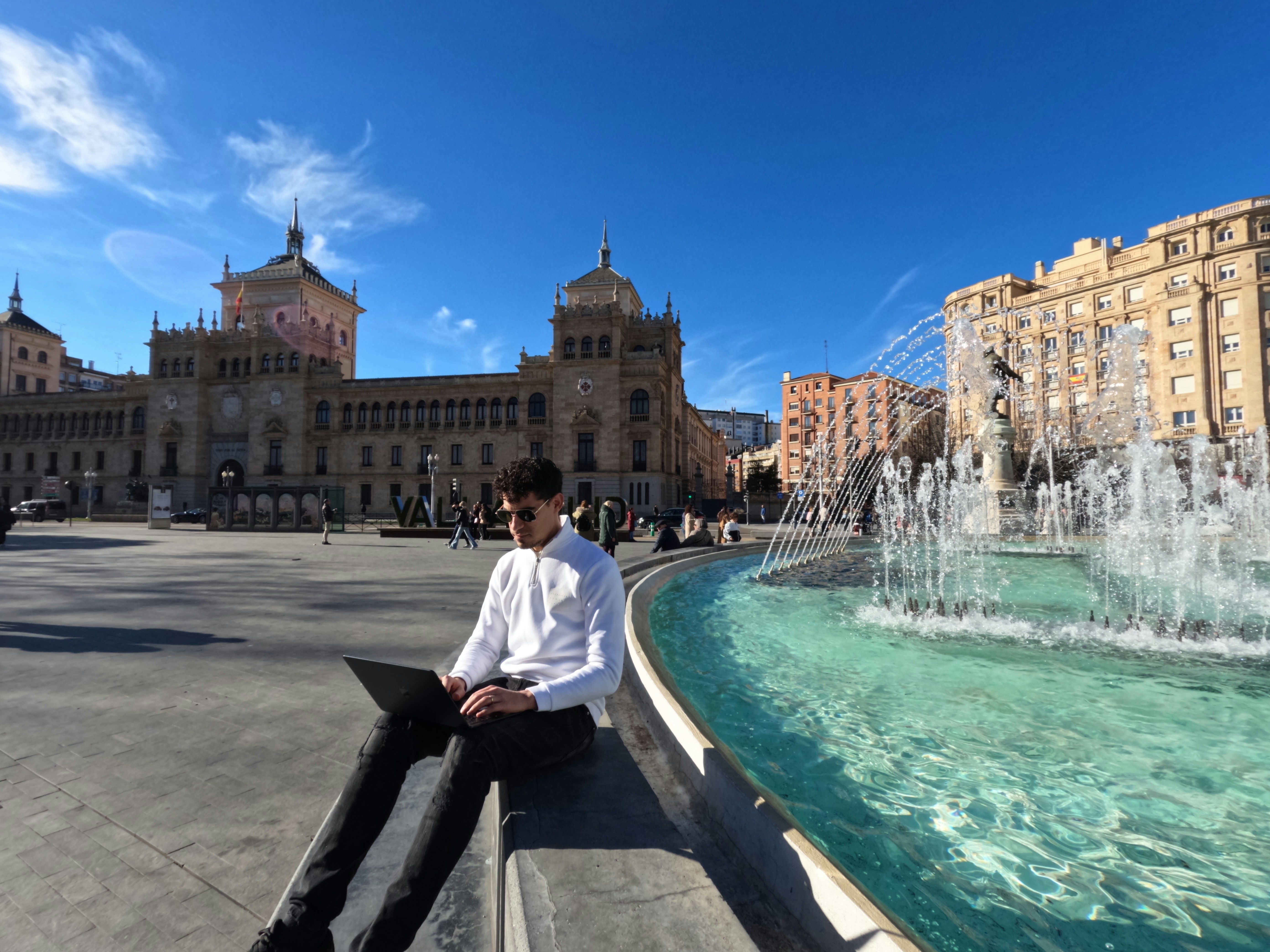 Hombre joven trabajando con un portátil al aire libre junto a una fuente en una plaza española.