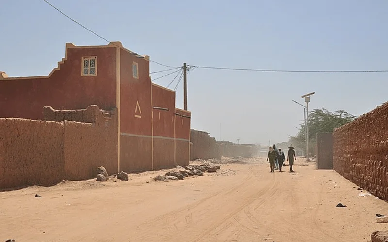 Vista de una calle polvorienta en In-Gall, con edificios de adobe rojizo, personas caminando y cielo azul claro en el desierto