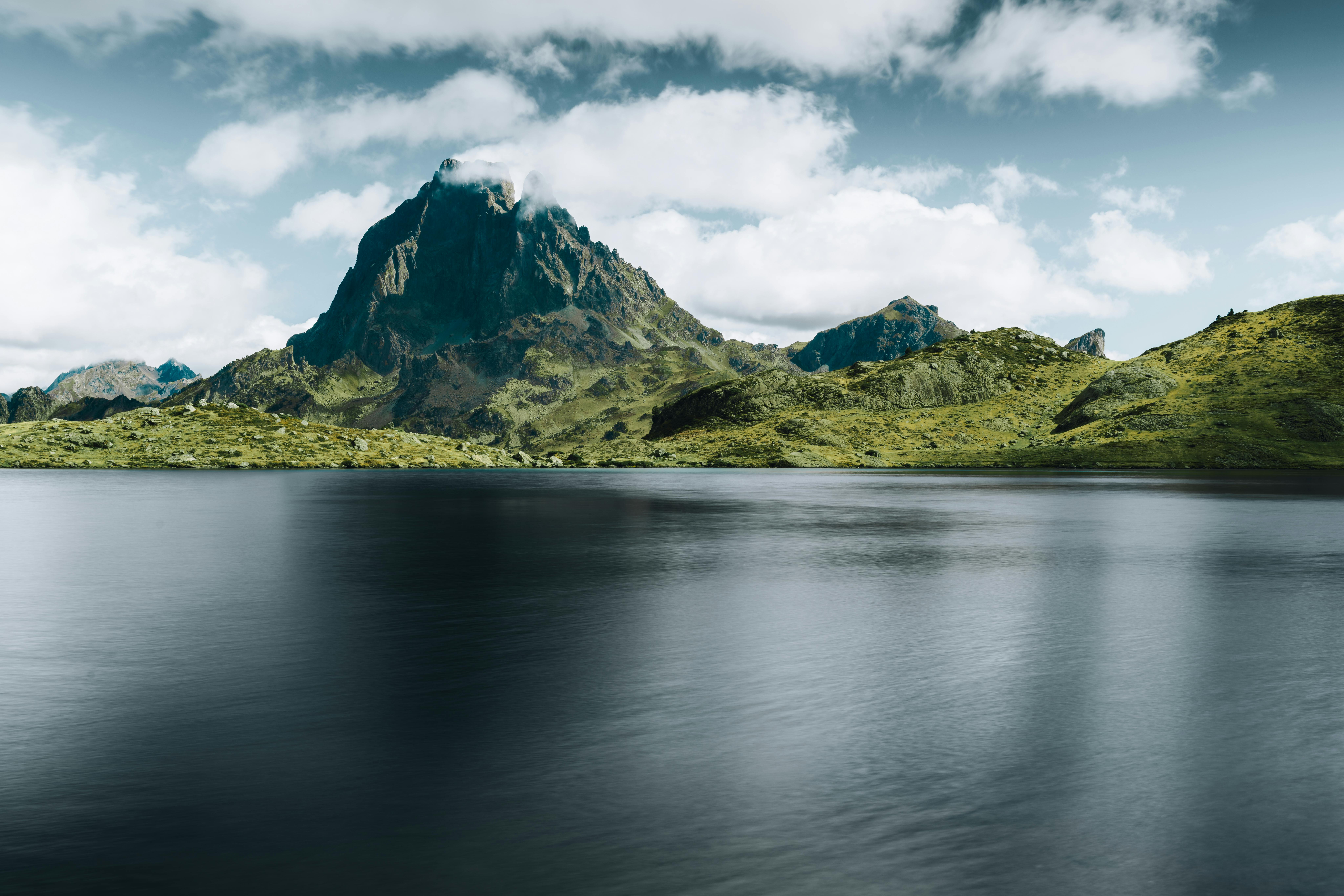 Lago de montaña con aguas oscuras y tranquilas, rodeado de colinas verdes y una gran montaña rocosa bajo nubes.