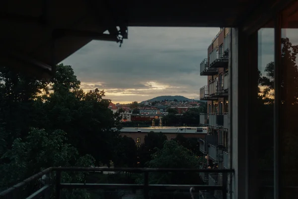 Vista de un paisaje urbano al atardecer desde un balcón, con edificios, árboles y una montaña al fondo.
