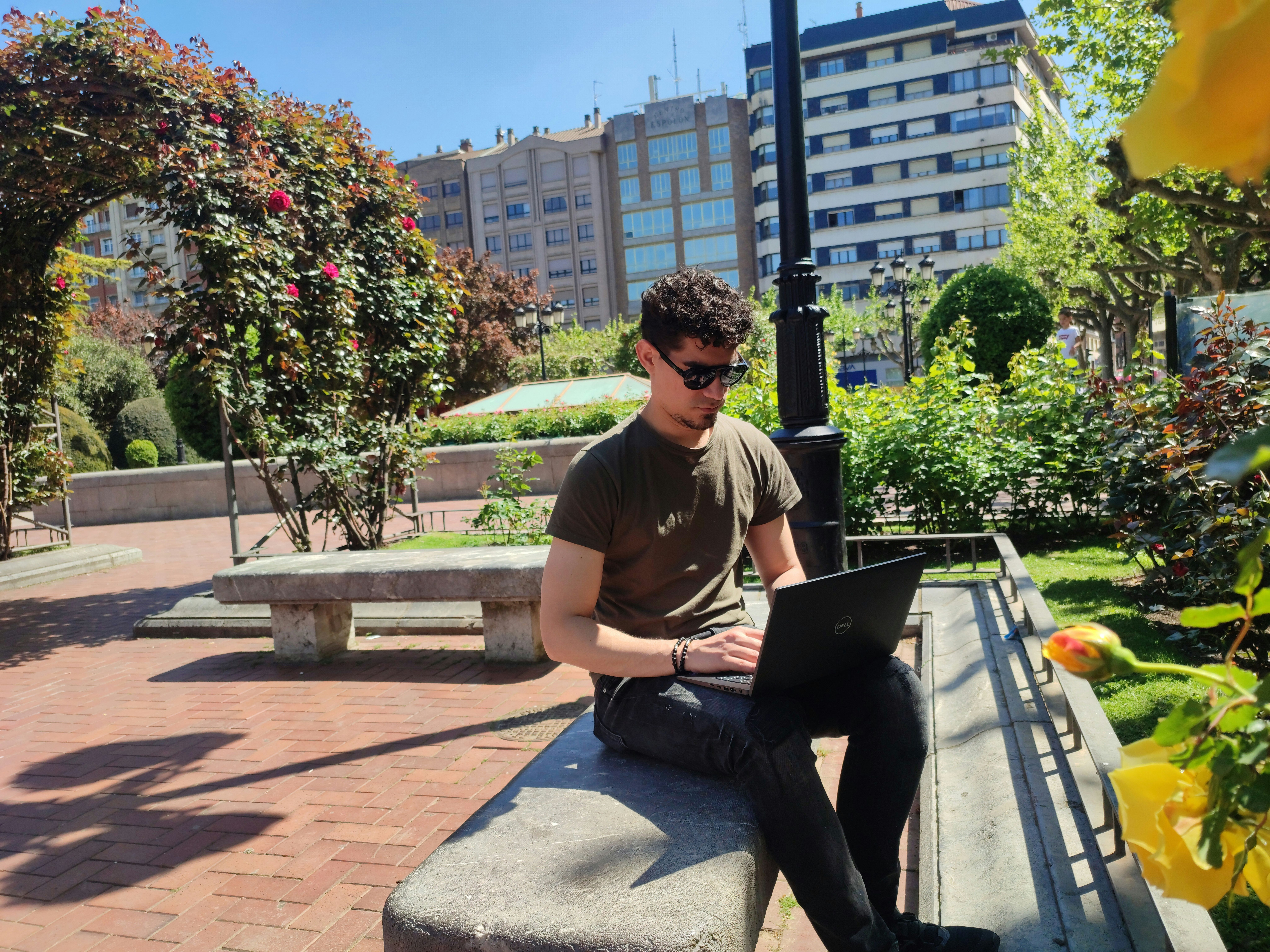 Hombre joven con gafas de sol trabajando en un portátil en un banco de piedra en un parque urbano.
