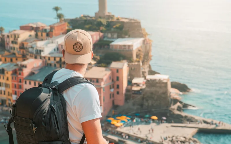 Hombre de espaldas con gorra y mochila observando un pueblo costero con casas coloridas y el mar.