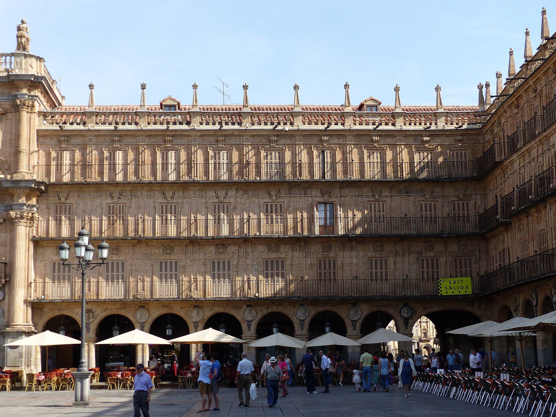 Vista de una plaza histórica con edificios de piedra de tres pisos, arcos y balcones, con gente y sombrillas.