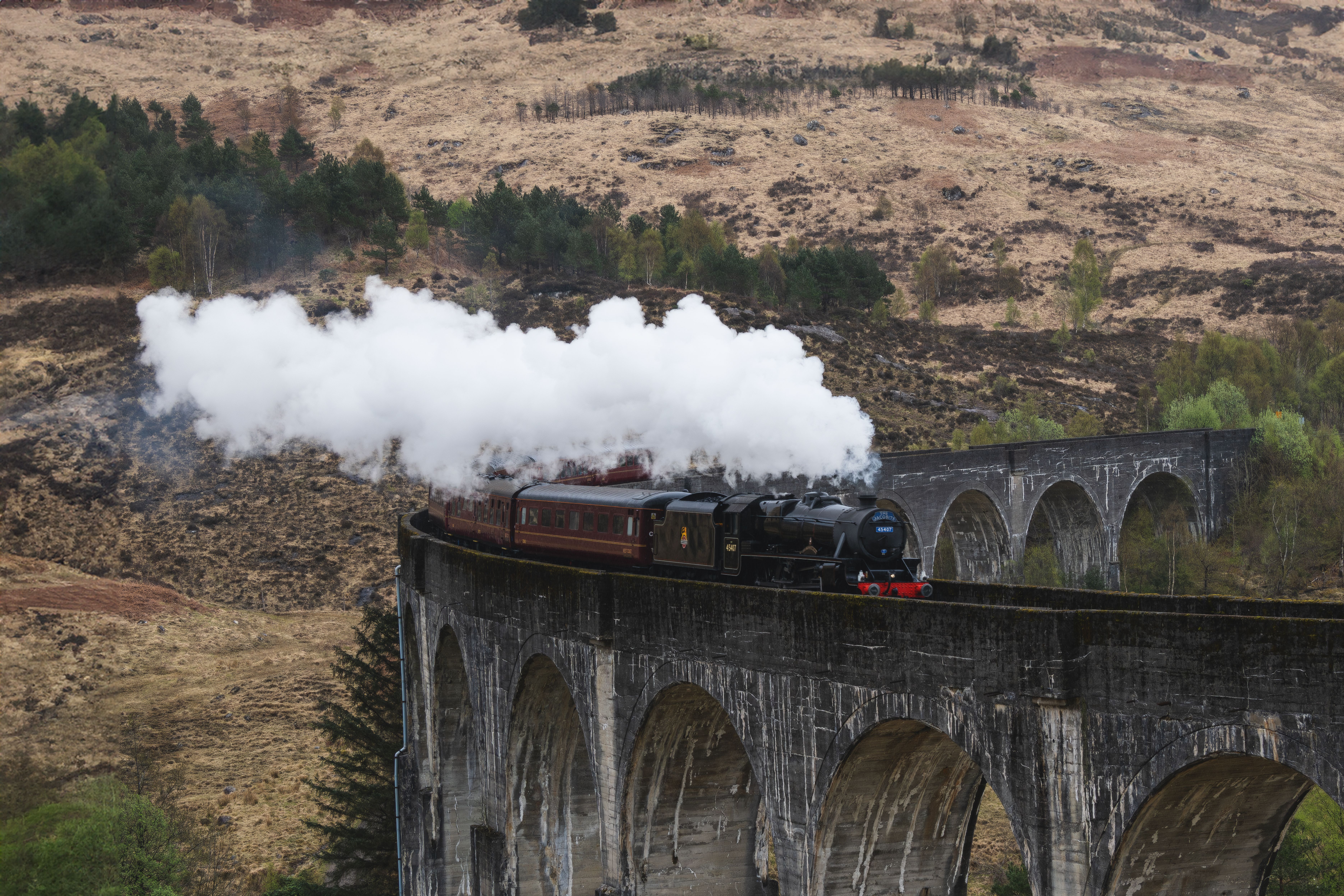 Tren de vapor negro y marrón cruzando un viaducto de piedra con arcos sobre un paisaje montañoso.