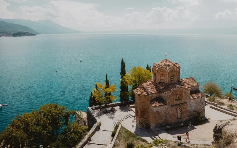 Vista aérea de una iglesia de piedra con tejado de tejas rojas junto a un gran lago azul.