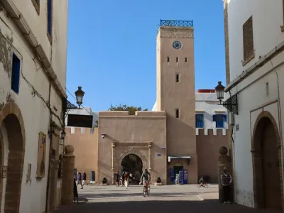 Vista de una calle empedrada en Essaouira con fachadas blancas, torre de reloj alta y personas caminando bajo cielo azul claro