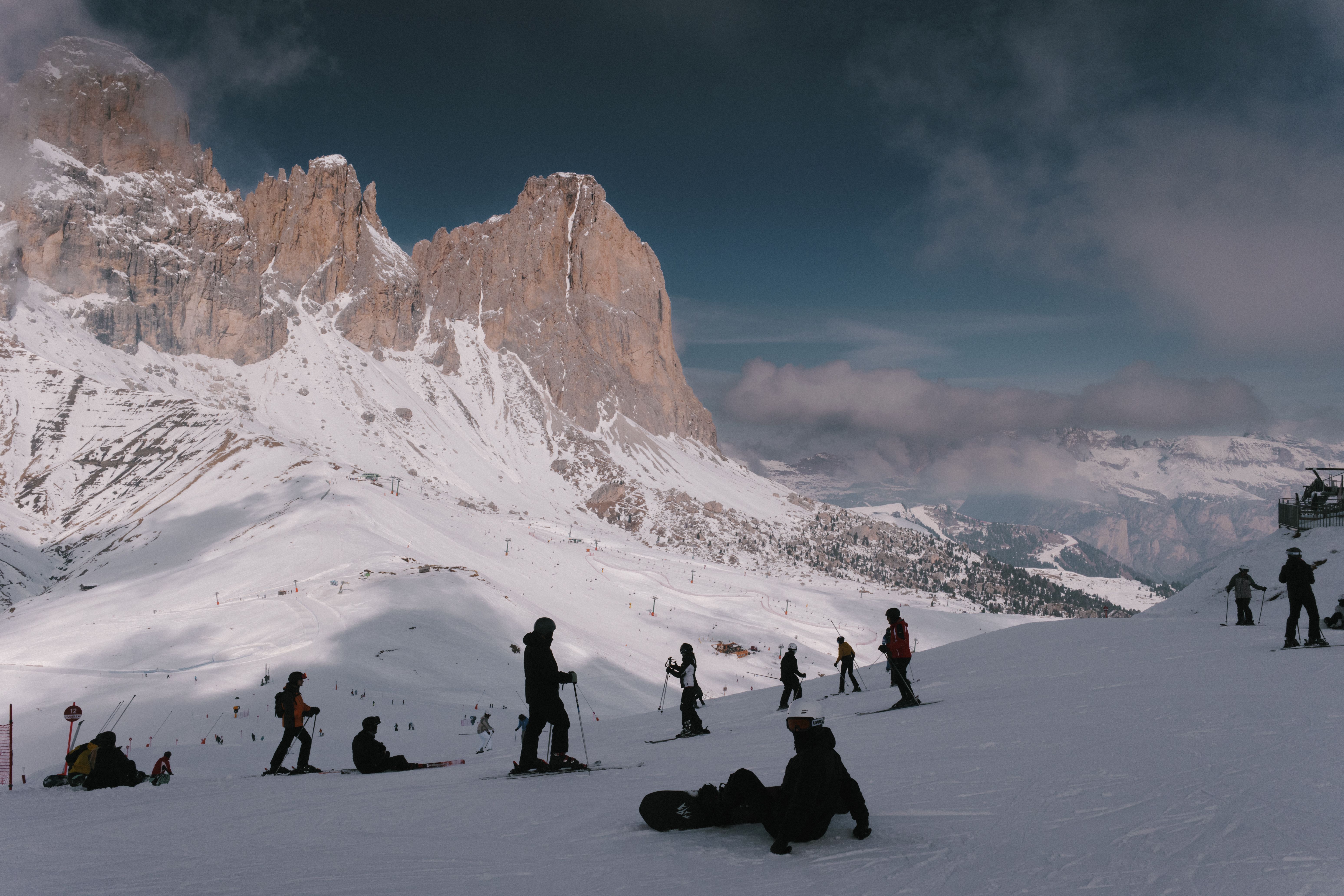 Esquiadores y snowboarders en una pista de nieve con montañas rocosas y cielo nublado de fondo.