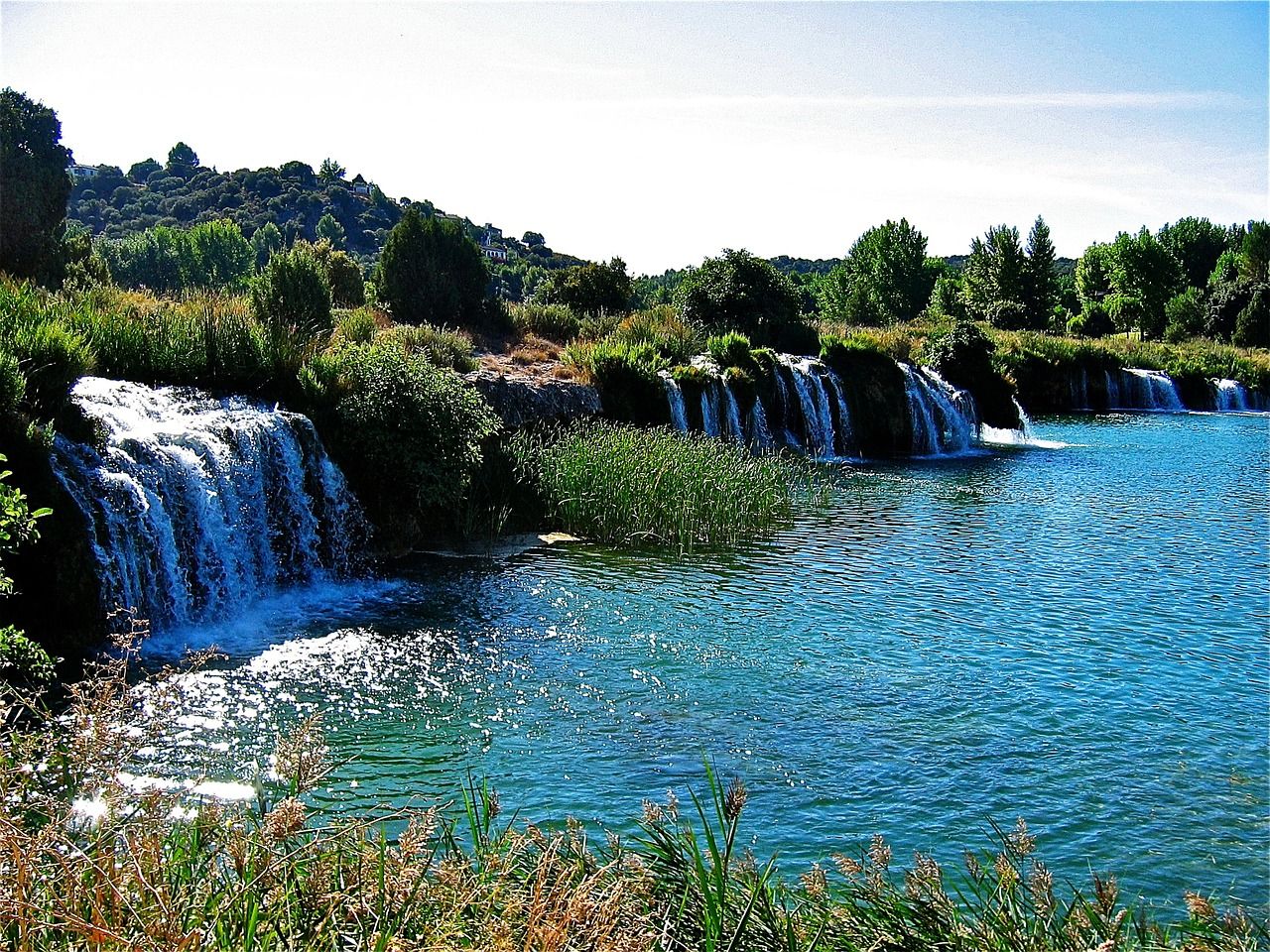 Cascadas de agua azul turquesa cayendo sobre rocas y vegetación verde bajo un cielo claro.