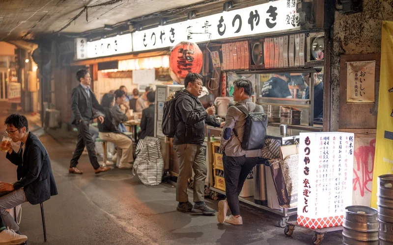 Hombres bebiendo y socializando en un callejón iluminado con letreros japoneses y faroles rojos.