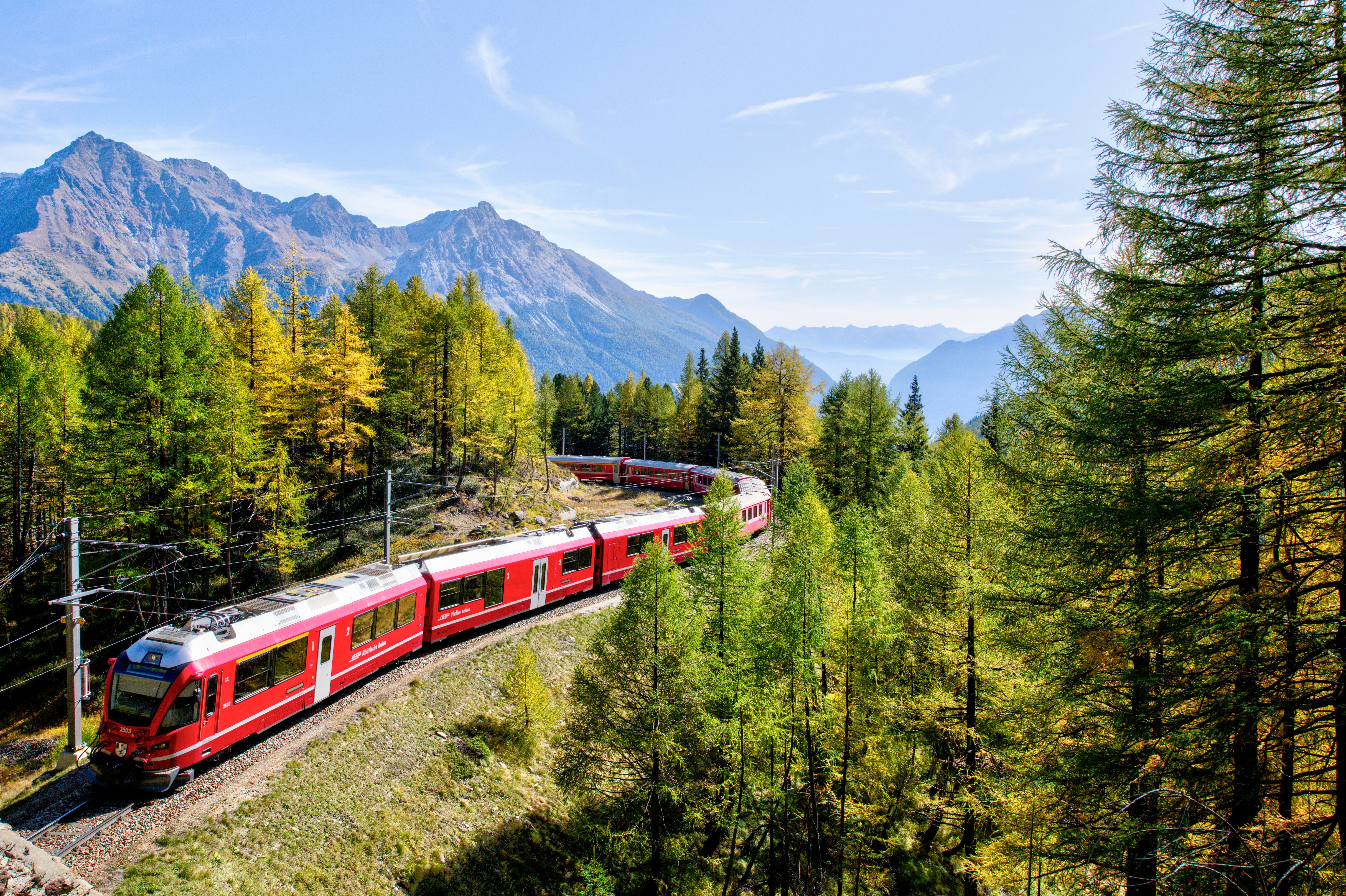 Tren rojo serpenteando por un valle montañoso cubierto de árboles verdes y amarillos bajo un cielo azul.