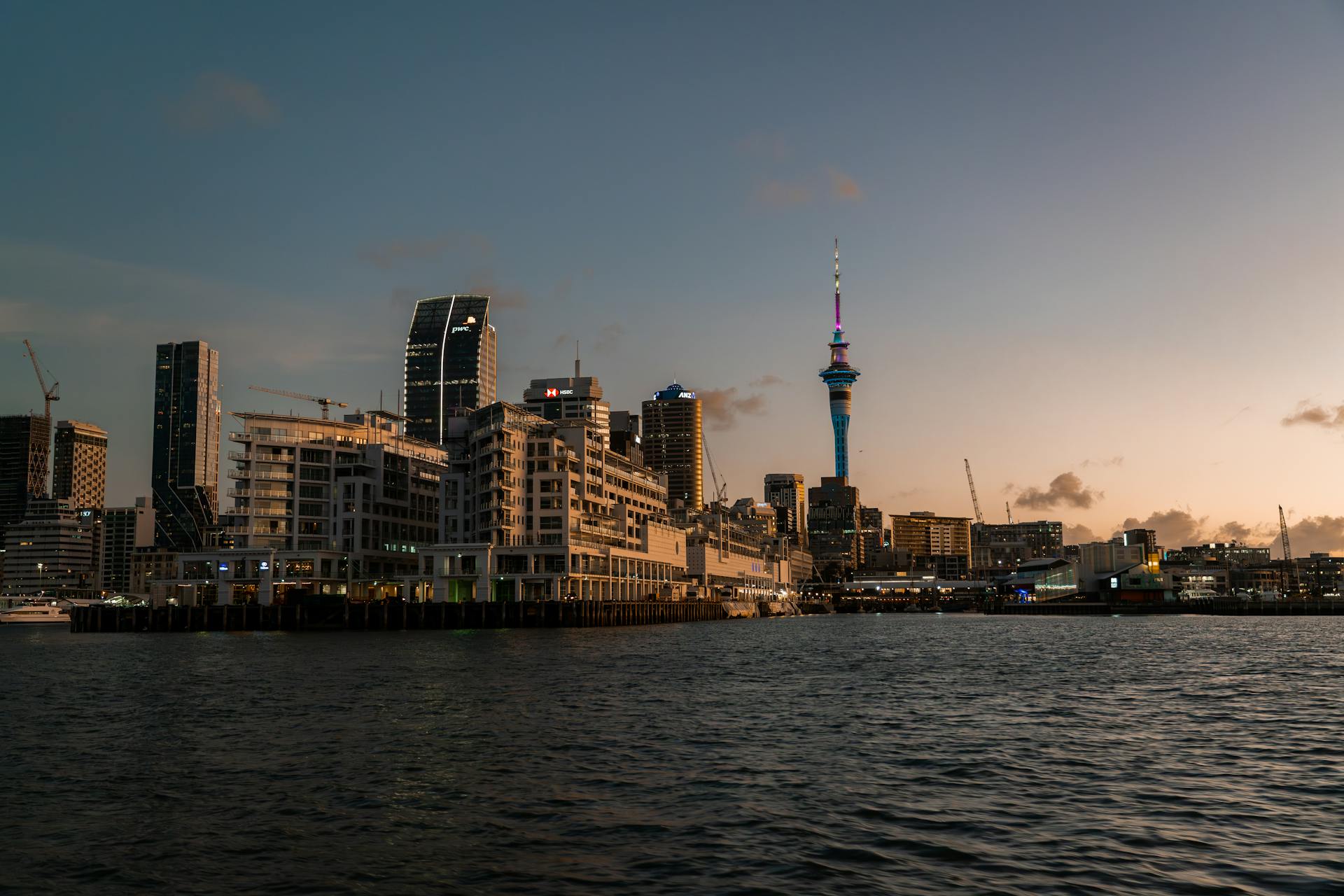 Panorama del skyline de Auckland al atardecer, con la torre Sky Tower iluminada en azul y edificios modernos junto al puerto