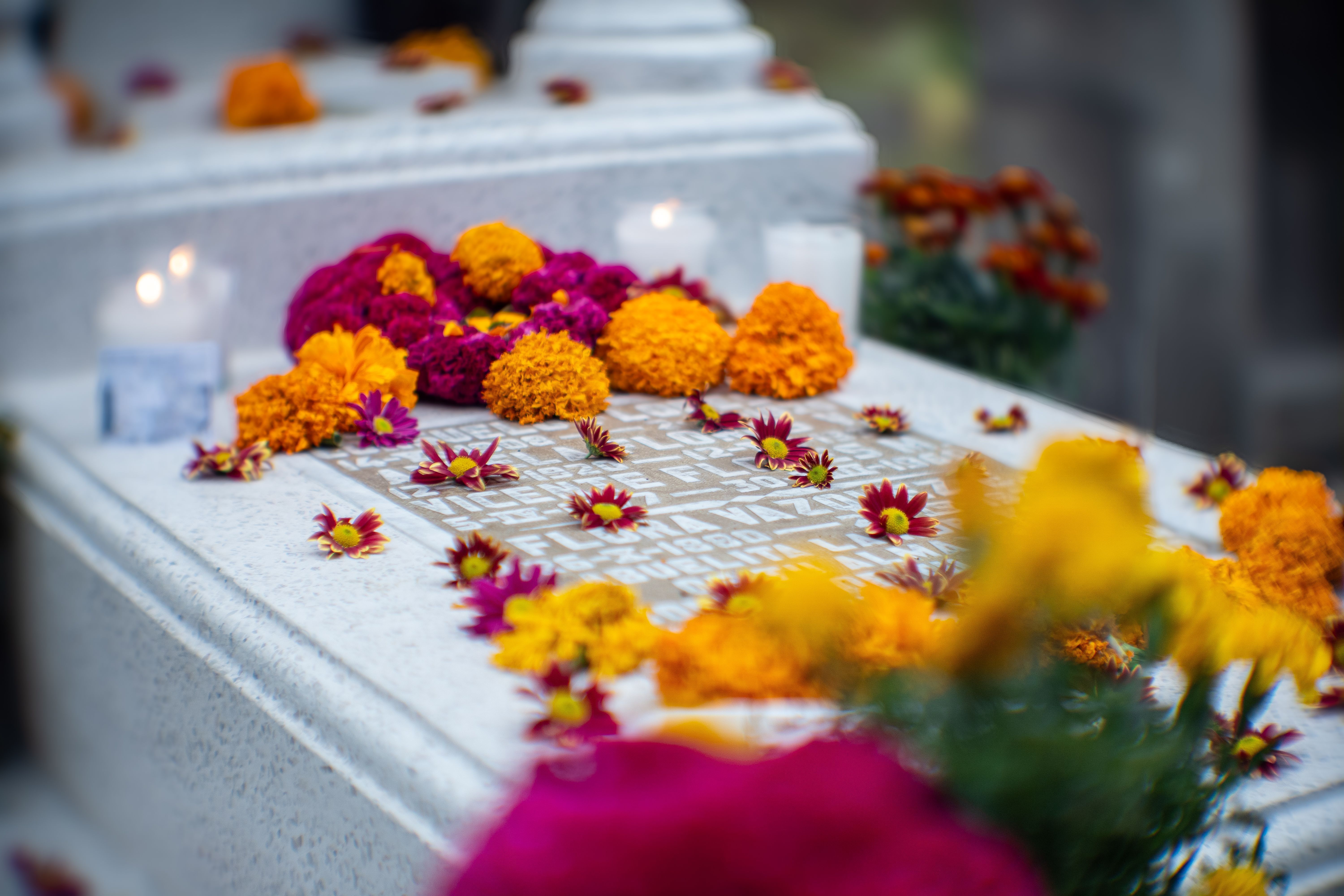Lápida de mármol blanco decorada con flores de cempasúchil naranjas, flores moradas y velas encendidas.