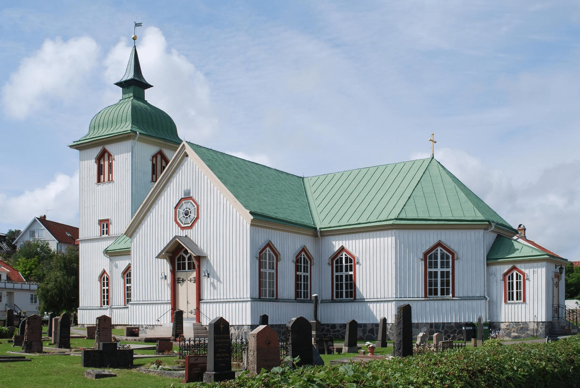 Iglesia de madera blanca con techo y cúpula verde, reloj en la fachada y detalles rojos, rodeada de un cementerio en un entorno rural soleado