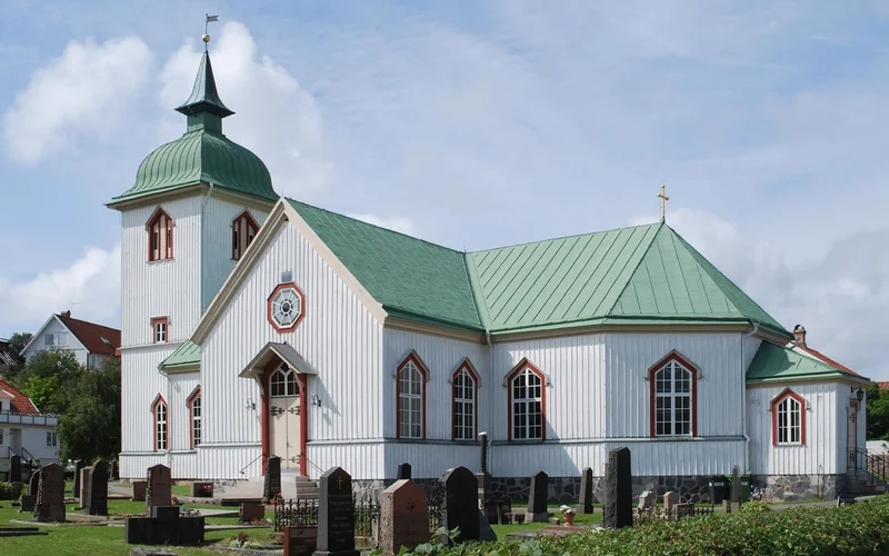 Iglesia de madera blanca con techo y cúpula verde, reloj en la fachada y detalles rojos, rodeada de un cementerio en un entorno rural soleado