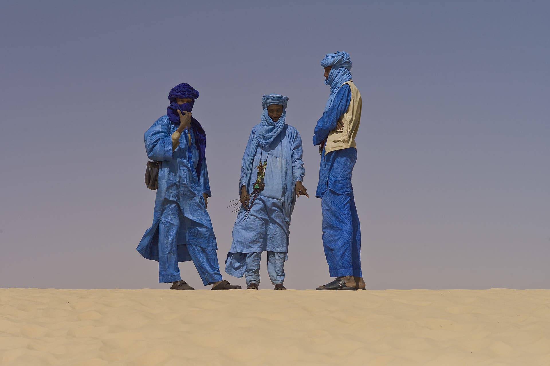 Tres hombres tuareg con túnicas azules indígenas posan en dunas doradas del desierto bajo un cielo azul claro