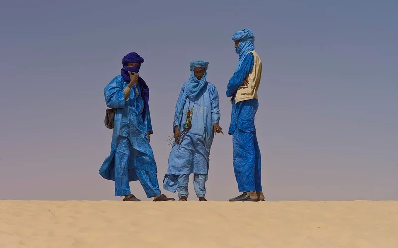 Tres hombres tuareg con túnicas azules indígenas posan en dunas doradas del desierto bajo un cielo azul claro