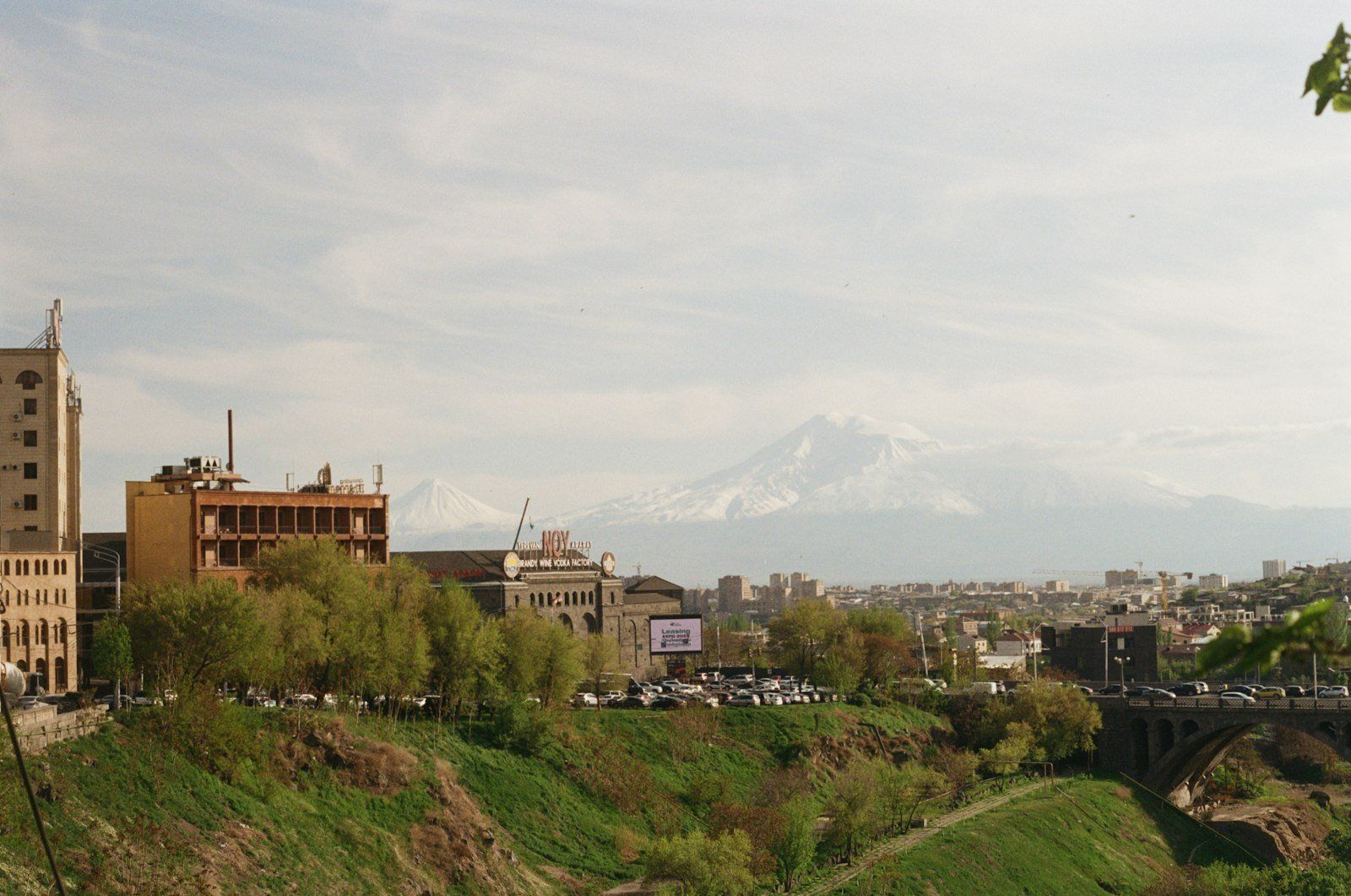 Panorama de la ciudad de Goris con edificios antiguos y modernos, monte Ararat nevado al fondo, vegetación verde y un puente sobre el río