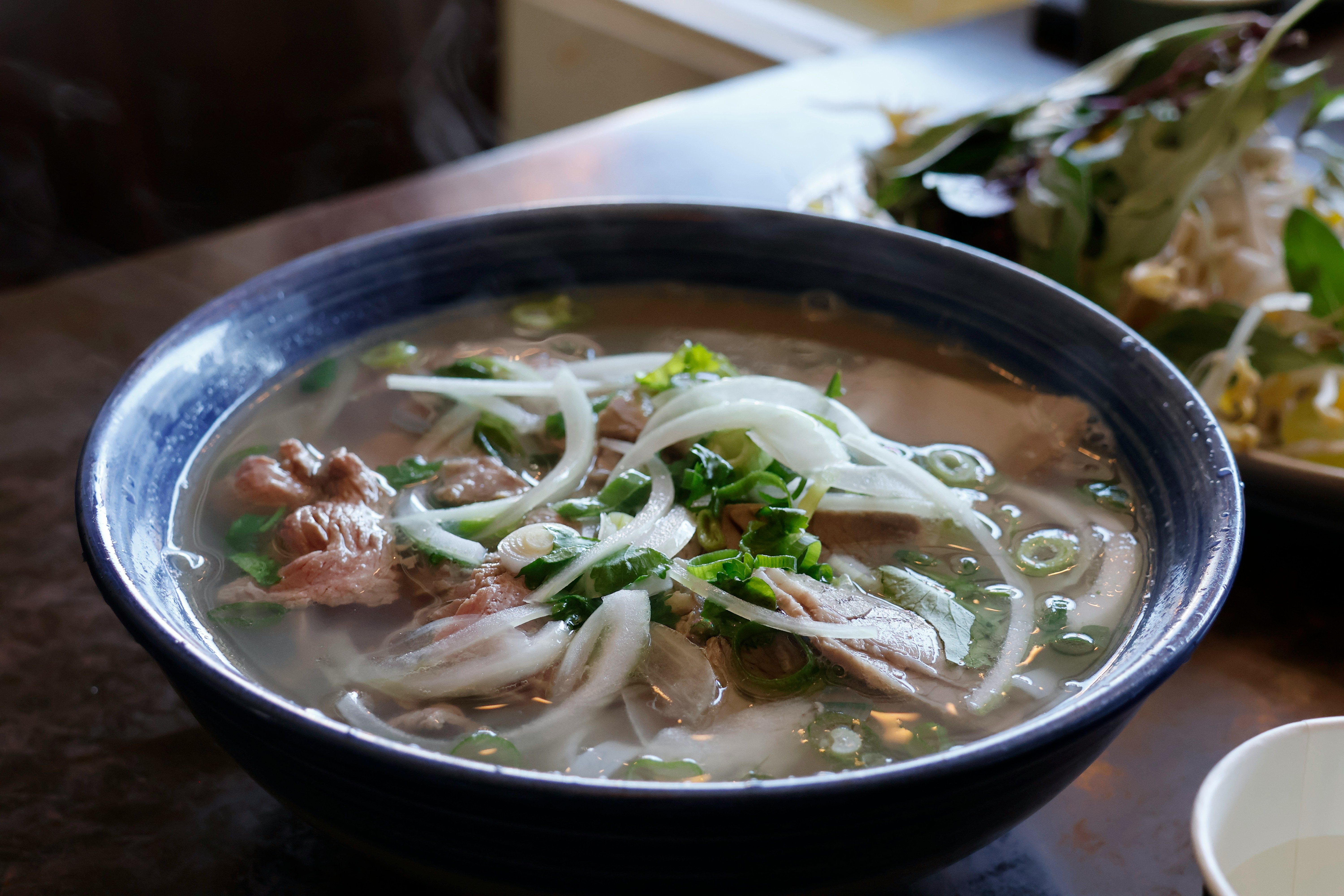 Un tazón de phở humeante con carne, cebolla y hierbas frescas sobre una mesa.