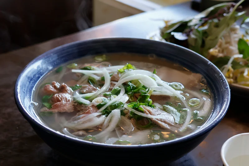 Un tazón de phở humeante con carne, cebolla y hierbas frescas sobre una mesa.