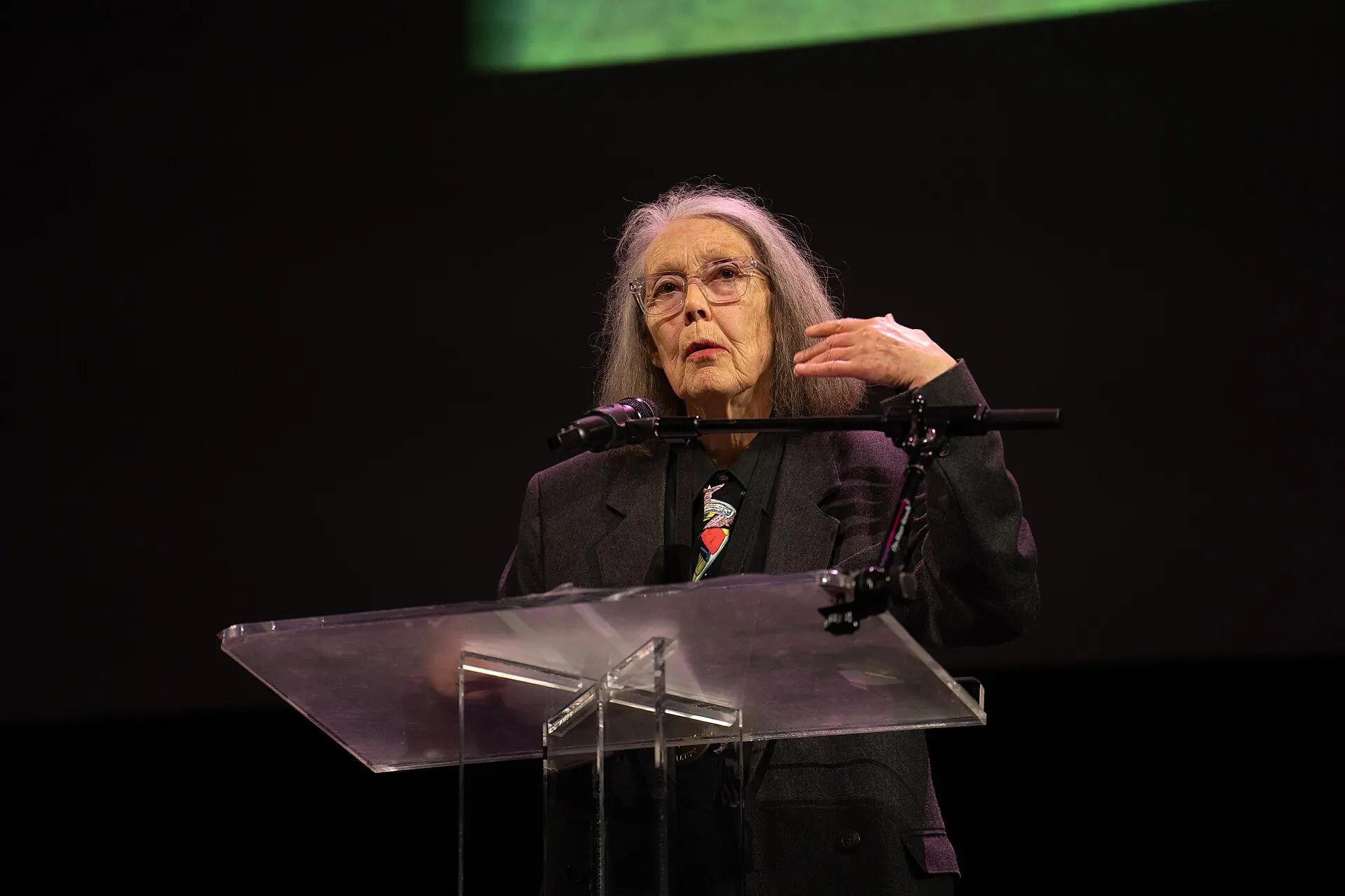 Anne Carson lee en un atril, con gafas y el pelo largo y canoso
