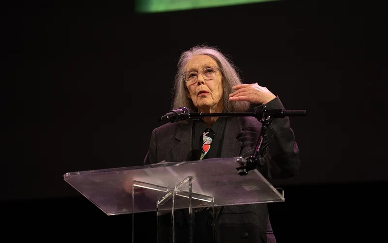 Anne Carson lee en un atril, con gafas y el pelo largo y canoso