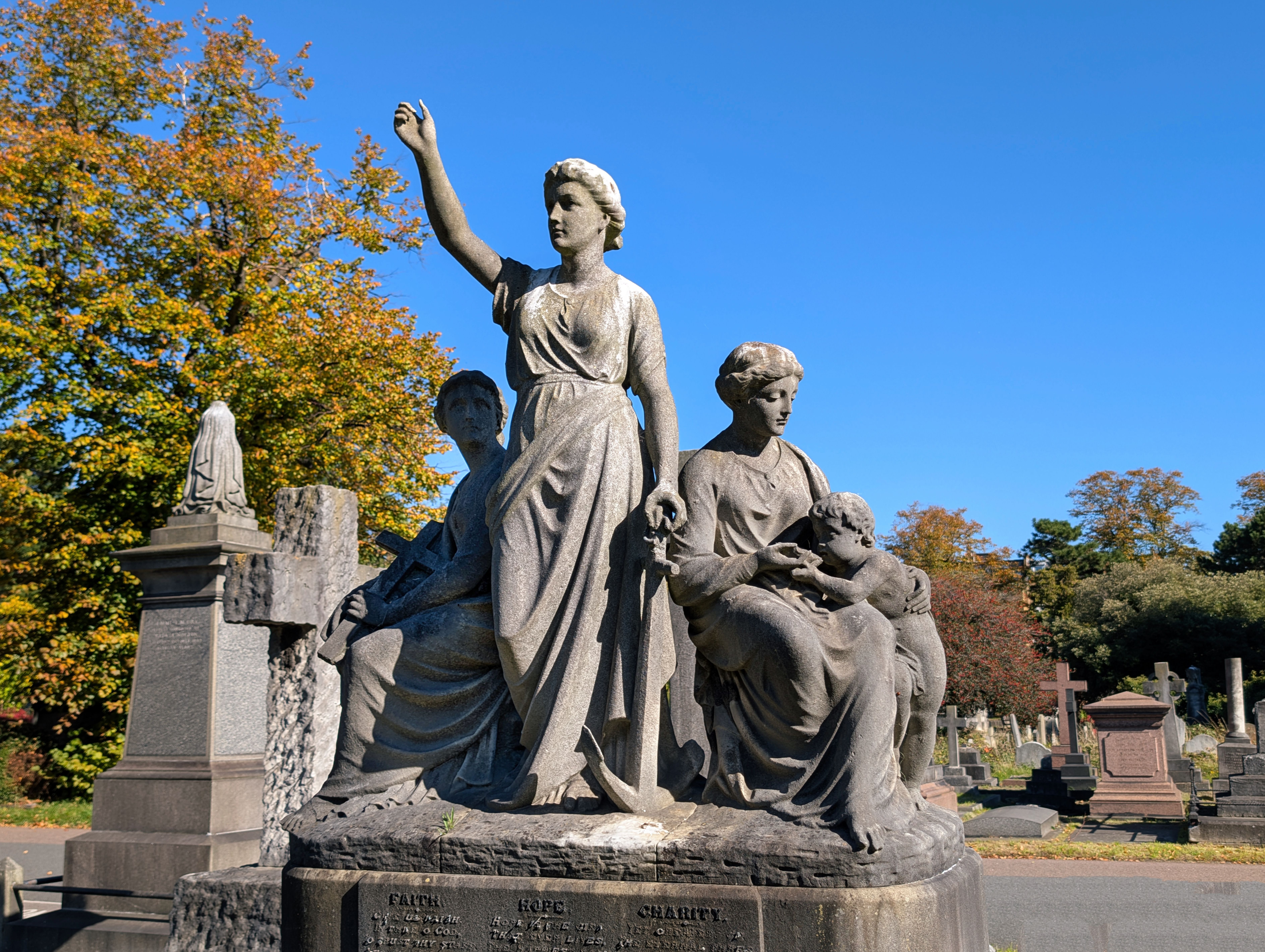 Escultura de piedra de tres figuras femeninas y un niño en un cementerio bajo un cielo azul.