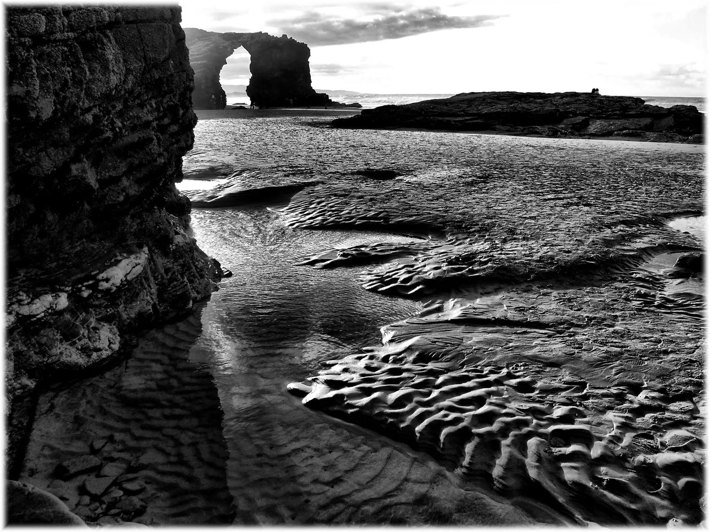 Vista en blanco y negro de la Playa de las Catedrales, con arcos rocosos dramáticos, arena ondulada y olas en la costa gallega