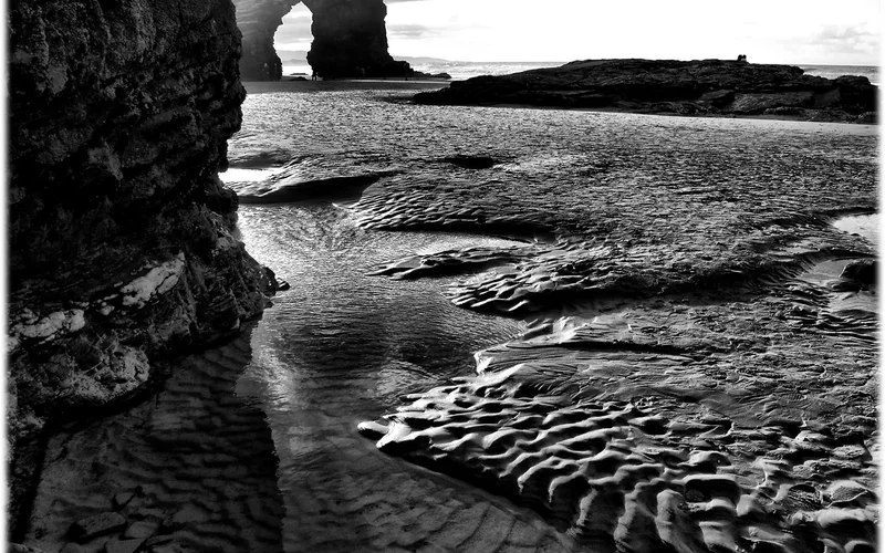 Vista en blanco y negro de la Playa de las Catedrales, con arcos rocosos dramáticos, arena ondulada y olas en la costa gallega
