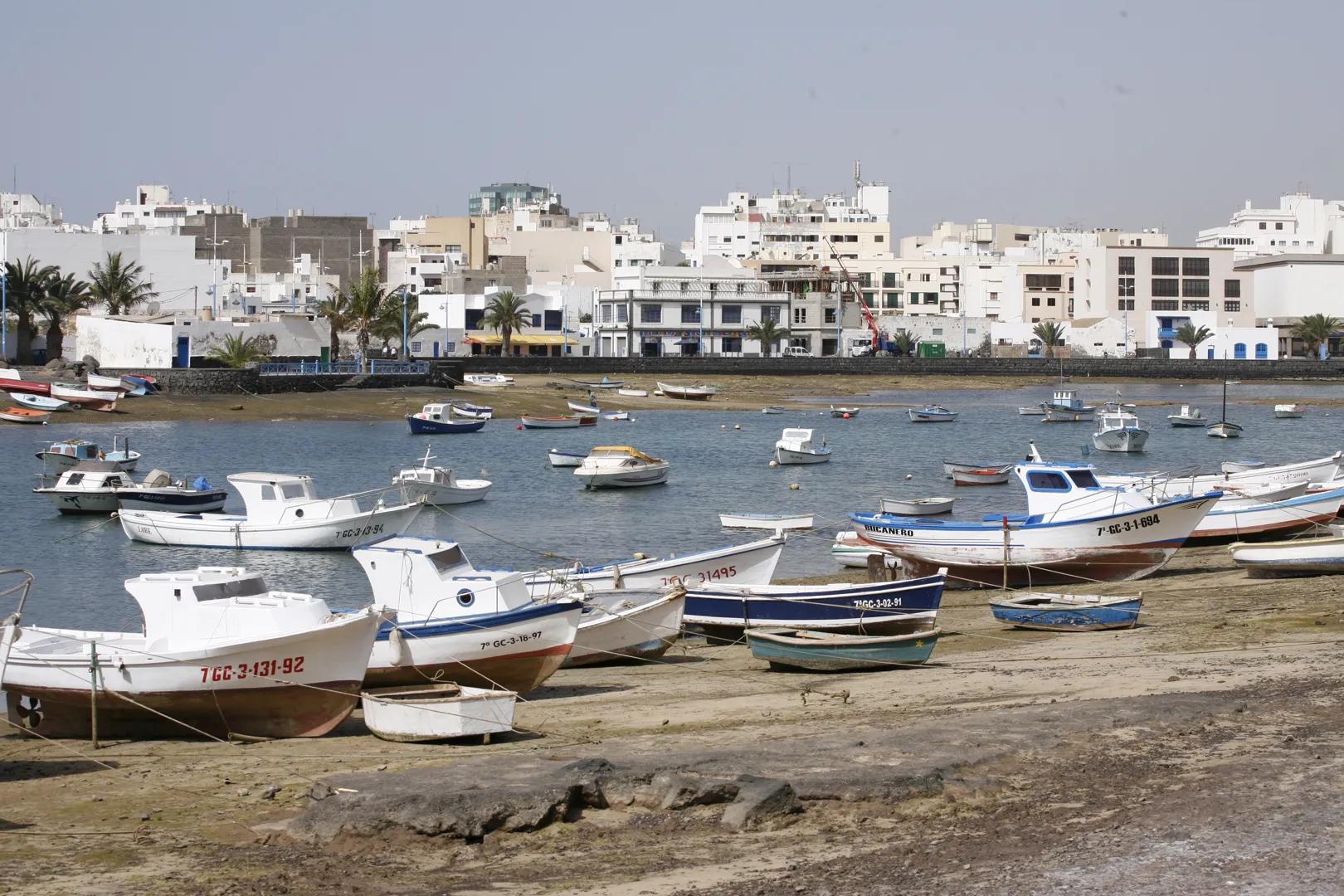 Vista de un puerto con barcos blancos y azules atracados en la orilla rocosa y flotando en el agua calma, edificios blancos y palmeras al fondo bajo