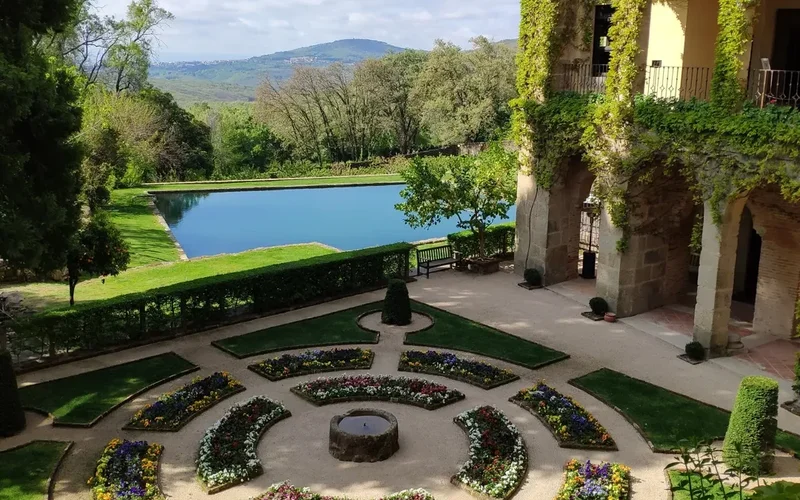 Vista de los jardines del Monasterio de Yuste con parterres florales circulares, piscina azul y edificio de piedra cubierto de hiedra, rodeado de
