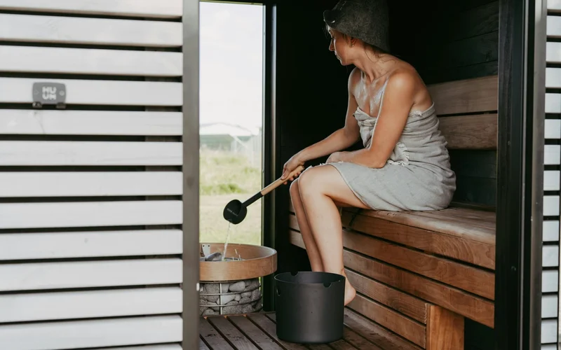 Una mujer en toalla sentada en un banco de madera dentro de una sauna, vertiendo agua.